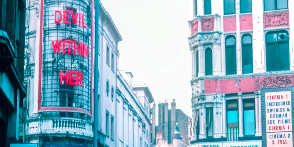 Street view of city buildings with neon sign reading "Devil Within Her" and adjacent historic architecture.