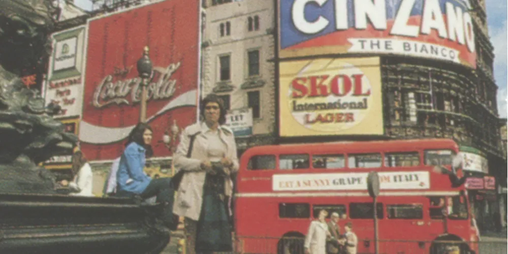 City street scene with red double-decker bus and iconic Coca-Cola and Cinzano billboards in a busy urban square.